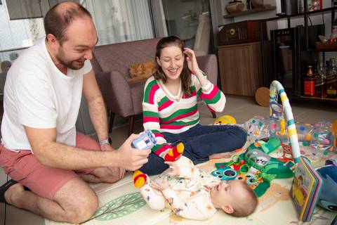 Pai e mãe sorrindo brincando com o bebê no tapetinho de atividades, em um momento feliz de interação familiar em ensaio de família em SP'