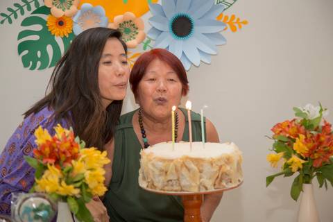 Aniversariante Reiko e sua filha assoprando as velas do bolo juntas, um momento icônico e emocionante da celebração do aniversário de 74 anos.'