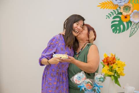 Aniversariante Reiko beijando a bochecha da filha com um sorriso, ao entregar-lhe o primeiro pedaço de bolo, em um momento alegre e emocionante.'