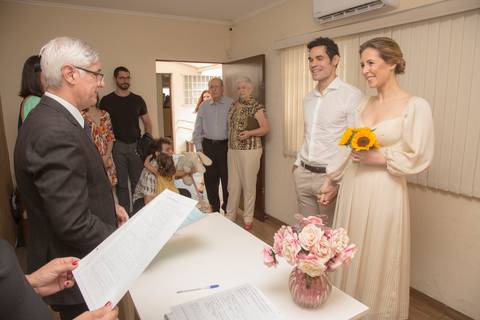 Juiz de paz falando para os noivos Ana e Leonardo, que estão sorridentes e atentos durante a cerimônia de casamento civil'
