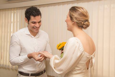 Noivo Leonardo colocando a aliança no dedo da noiva Ana, que sorri emocionada durante a troca de alianças no cartório'