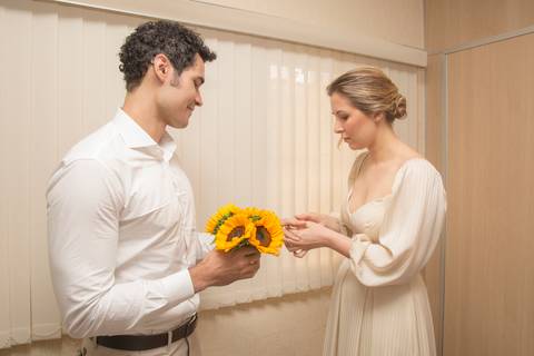 Noiva Ana colocando a aliança no dedo do noivo Leonardo, que sorri, selando a união no casamento civil'