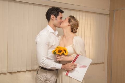 Casal Ana e Leonardo se beijando carinhosamente, segurando a certidão de casamento em mãos, celebrando a união no cartório'