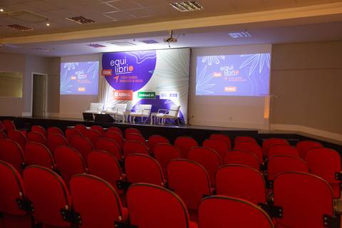 Espaço do Fórum Equilíbrio da ABRH SC, com fileiras de cadeiras vermelhas, palco iluminado ao fundo e banner de identificação, com cadeiras para os palestrantes. Fotografia Bruno Arita'