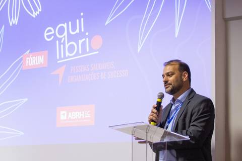 Abertura do Fórum Equilíbrio da ABRH SC: organizador discursando no palco, com a identidade visual do evento projetada no telão ao fundo. Fotografia Bruno Arita'