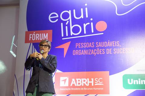 Mulher palestrante falando no palco do Fórum Equilíbrio da ABRH SC, com o painel do evento ao fundo, em um momento de apresentação. Fotografia Bruno Arita'