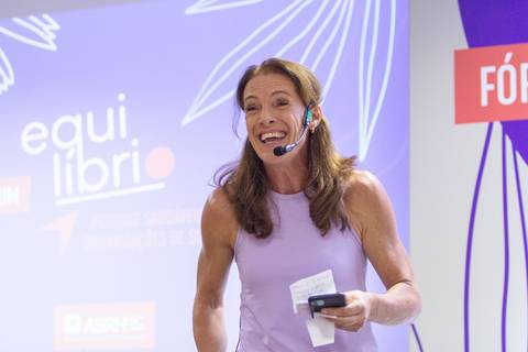 Ex-atleta palestrante falando e sorrindo no palco do Fórum Equilíbrio da ABRH SC, com a identidade visual do evento ao fundo. Fotografia Bruno Arita'
