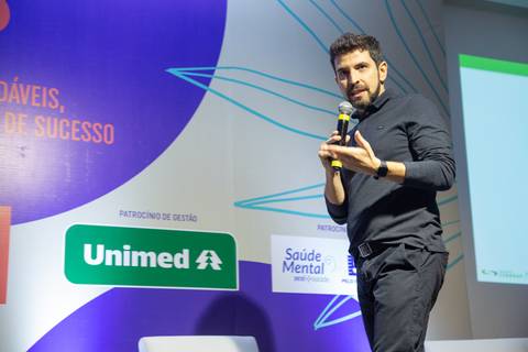 Médico palestrante falando no palco do Fórum Equilíbrio da ABRH SC, com a identidade visual do evento projetada por trás dele. Fotografia Bruno Arita'