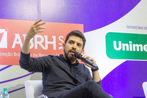Plano fechado no médico palestrante do Fórum Equilíbrio da ABRH SC, sentado em uma cadeira no palco e falando ao público. Fotografia Bruno Arita'
