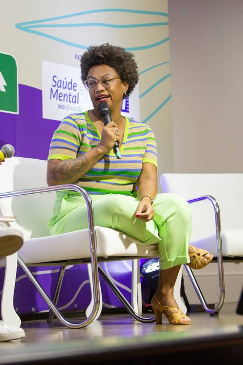 Mediadora sentada no palco do Fórum Equilíbrio da ABRH SC, falando no microfone em plano fechado, conduzindo a discussão. Fotografia Bruno Arita'