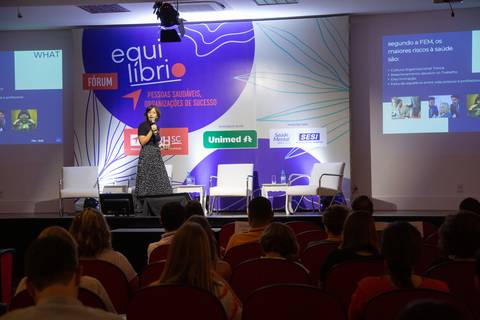 Plano aberto do Fórum Equilíbrio da ABRH SC, mostrando parte do público e a palestrante no palco, com o banner do evento visível ao fundo. Fotografia Bruno Arita'