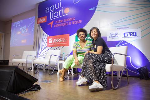 Mediadora e palestrante posando para foto sentadas em cadeiras no palco do Fórum Equilíbrio da ABRH SC, com a identidade visual do evento ao fundo. Fotografia Bruno Arita'