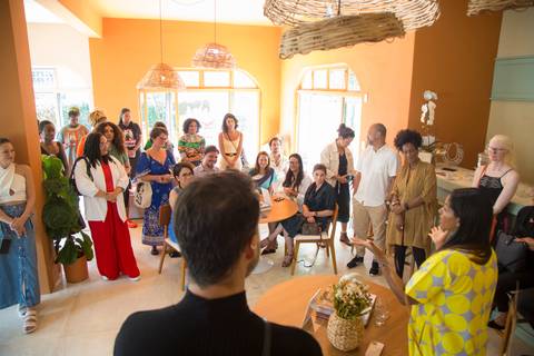 Plano aberto de Alexandra Loras discursando para convidados que a ouvem atentos ao fundo, mostrando a amplitude do evento de inauguração'