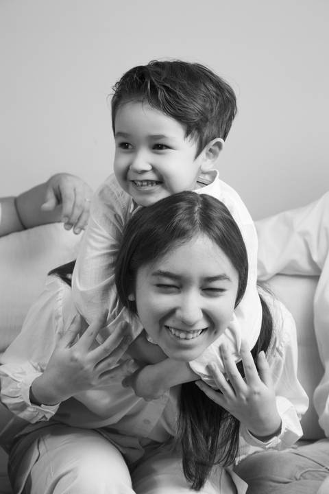 Filho de 5 anos fazendo cavalinho na irmã adolescente, ambos rindo em uma foto divertida em preto e branco, capturando a cumplicidade entre irmãos. (Ensaio de Família, São Paulo, Fotógrafo Bruno Arita)'