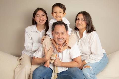 Família (pai, mãe, filha adolescente e filho de 5 anos) sentada no encosto do sofá, com o filho de 5 anos no pescoço do pai, em pose divertida e conectada. (Ensaio de Família, São Paulo, Fotógrafo Bruno Arita)'