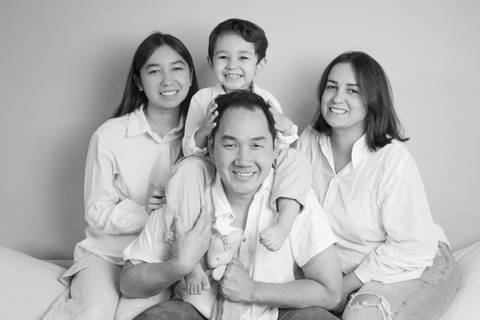Fotografia em preto e branco de filha adolescente, filho de 5 anos sentado de cavalinho no pescoço do pai, e mãe, sentados juntos no encosto alto do sofá, em momento de alegria familiar. (Ensaio de Família, São Paulo, Fotógrafo Bruno Arita)'