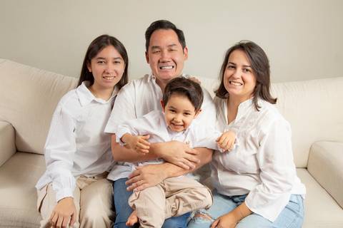 Filha adolescente com camisa branca e calça creme, pai de camisa social branca e jeans, filho de 5 anos no colo do pai, e mãe de camisa branca e jeans, todos sentados juntos no sofá branco, transmitindo união. (Ensaio de Família, São Paulo, Fotógrafo Brun'