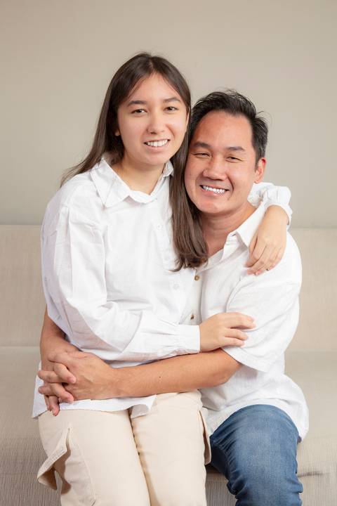 Filha adolescente e pai sentados no sofá, posando para a foto com um sorriso, transmitindo a conexão entre eles. (Ensaio de Família, São Paulo, Fotógrafo Bruno Arita)'