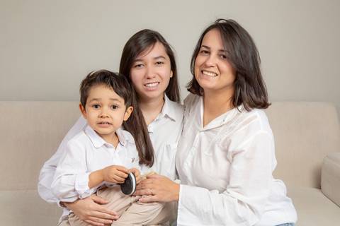 Filho de 5 anos no colo da irmã adolescente, ao lado da mãe (camisa branca e jeans), todos sentados juntos no sofá branco, em um registro de conexão familiar. (Ensaio de Família, São Paulo, Fotógrafo Bruno Arita)'