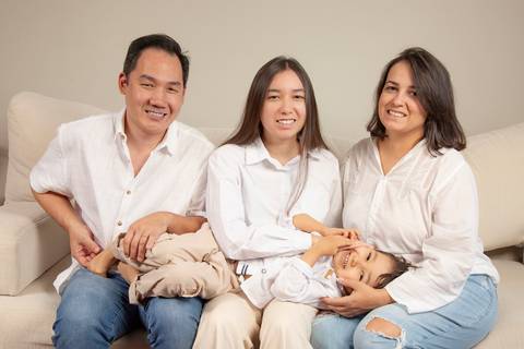 Pai, filha adolescente e mãe (todos de camisa branca/creme e jeans), sentados no sofá, com o filho de 5 anos deitado confortavelmente no colo dos três, em um momento de união e aconchego. (Ensaio de Família, São Paulo, Fotógrafo Bruno Arita)'