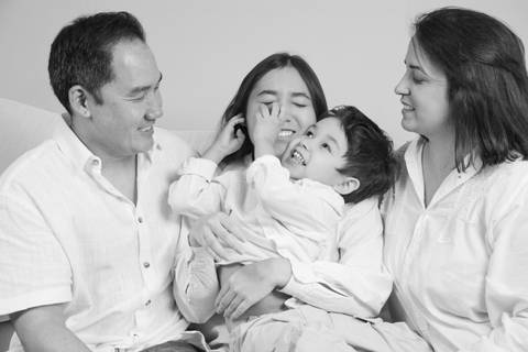 Fotografia em preto e branco da filha adolescente sentada entre o pai e a mãe, com o irmão de 5 anos no colo dela, mexendo carinhosamente no rosto da irmã em um momento de brincadeira espontânea. (Ensaio de Família, São Paulo, Fotógrafo Bruno Arita)'