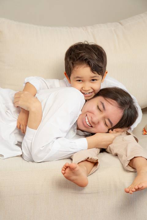 Filha adolescente deitada com os olhos fechados, sorrindo, no colo do irmão de 5 anos, que está sentado no sofá, em uma foto que irradia ternura entre os irmãos. (Ensaio de Família, São Paulo, Fotógrafo Bruno Arita)'