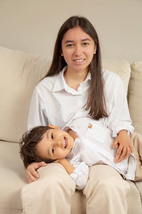 Filha adolescente (camisa branca e calça creme) com o irmão de 5 anos em seu colo, ambos com sorrisos e olhares de ternura, sentados no sofá branco. (Ensaio de Família, São Paulo, Fotógrafo Bruno Arita)'