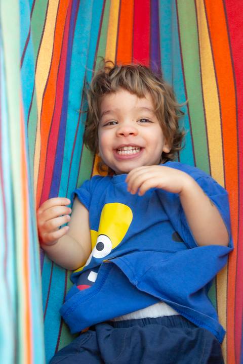 Criança sorrindo para a câmera, deitada em uma rede de descanso com listras coloridas em arco-íris, em plano fechado, transmitindo alegria. (Ensaio de Família, São Paulo, Fotógrafo Bruno Arita)'