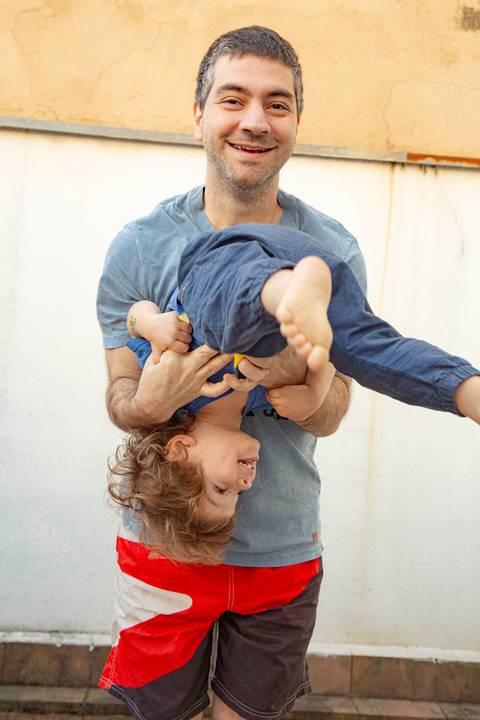 Pai de pé, carregando o filho pequeno de ponta cabeça no colo, em um momento de intensa brincadeira e alegria familiar. (Ensaio de Família, São Paulo, Fotógrafo Bruno Arita)'