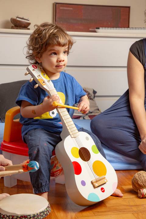 Criança pequena 'tocando' um violão infantil como se fosse um violoncelo, em um momento de fofura e brincadeira. (Ensaio de Família, São Paulo, Fotógrafo Bruno Arita)'