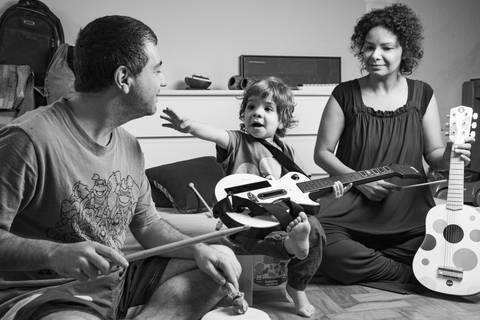 Foto em preto e branco da família brincando no chão: pai tocando tambor, filho pequeno no meio com uma guitarra de videogame acenando para o pai, e mãe segurando um violão infantil como um violoncelo. (Ensaio de Família, São Paulo, Fotógrafo Bruno Arita)'