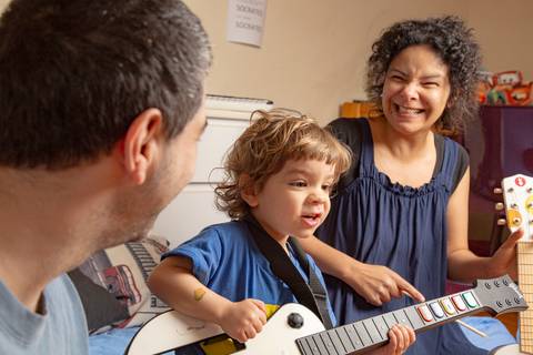 Plano fechado com pai observando o filho pequeno 'tocar' a guitarra de videogame, e a mãe sorrindo ao fundo, acompanhando a cena divertida. (Ensaio de Família, São Paulo, Fotógrafo Bruno Arita)'