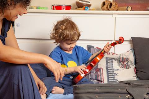Criança pequena recebendo um violino de presente, com o olhar carinhoso da mãe observando o momento. (Ensaio de Família, São Paulo, Fotógrafo Bruno Arita)'