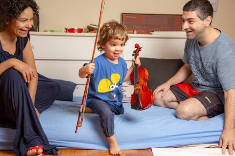 Criança pequena feliz segurando o violino de presente e a vareta, sob o olhar de alegria dos pais, todos juntos no colchão de brincadeira. (Ensaio de Família, São Paulo, Fotógrafo Bruno Arita)'