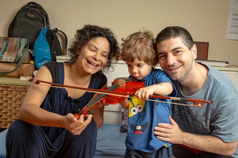 Família posando para foto: mãe observando o filho pequeno tentando tocar o violino e o pai sorrindo para a câmera, em um registro de união e apoio. (Ensaio de Família, São Paulo, Fotógrafo Bruno Arita)'