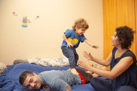 Pai deitado de bruços no colchão, com o filho pequeno andando em suas costas, prestes a pular em cima da mãe, em um momento de brincadeira e afeto. (Ensaio de Família, São Paulo, Fotógrafo Bruno Arita)'