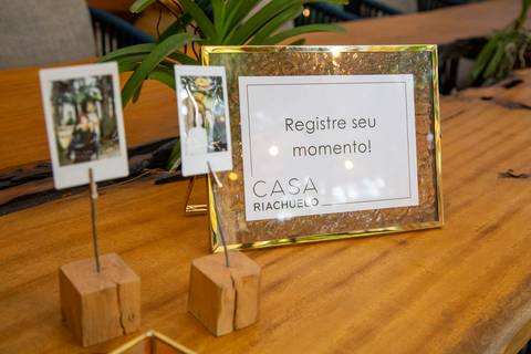 Mesa com duas amostras de fotos instantâneas presas por suportes que os convidados puderam levar, e um porta-retrato com a frase 'Registre seu momento' e o logo da Casa Riachuelo. (Evento Casa Riachuelo CASACOR 2025, São Paulo, Fotos Bruno Arita)'