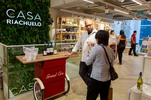Sommelier da Grand Cru conversando com uma cliente, em plano aberto, em frente ao carrinho de vinhos com o letreiro da Casa Riachuelo ao fundo. (Lançamento Casa Riachuelo, Rua Oscar Freire, São Paulo, Fotos Bruno Arita'