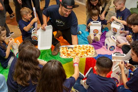 Recreador servindo lanche da tarde para as crianças, que estão sentadas no chão sobre panos coloridos, em um momento de pausa. (Evento Plurix, São Paulo, Fotógrafo Bruno Arita)'