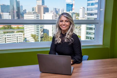 Retrato ambiental de Mulher sentada em sua mesa, olhando para a câmera com um notebook, em frente a uma janela com vista da cidade de São Paulo em dia ensolarado. (Retrato Corporativo, São Paulo, Fotógrafo Bruno Arita)'