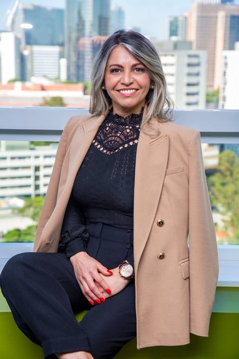 Retrato ambiental de Mulher sentada em frente a uma janela com vista de São Paulo, em dia ensolarado, com blazer claro apoiado nos ombros, em pose descontraída. (Retrato Corporativo, São Paulo, Fotógrafo Bruno Arita)'