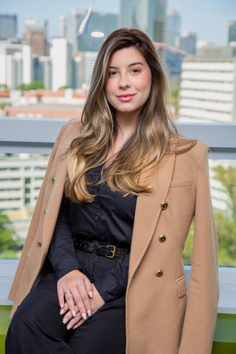 Retrato ambiental de Mulher sentada em pose elegante, com blazer claro nos ombros, em frente a uma janela com vista de São Paulo, com olhar sério e concentrado. (Retrato Corporativo, São Paulo, Fotógrafo Bruno Arita)'