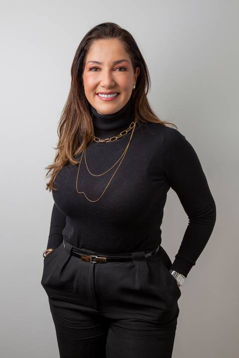 Retrato profissional de Mulher alta (cabelo castanho) de pé, em postura elegante, com mãos nos bolsos e sorrindo. Traje preto com colar dourado fino. (Retrato Corporativo, São Paulo, Fotógrafo Bruno Arita)'