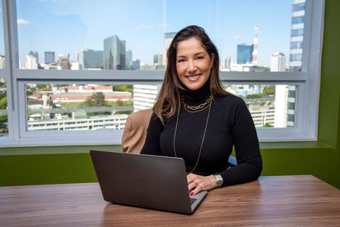 Retrato ambiental de Mulher sentada em mesa de trabalho com notebook, olhando e sorrindo para a câmera, com o skyline de São Paulo visível ao fundo. (Retrato Corporativo, São Paulo, Fotógrafo Bruno Arita)'