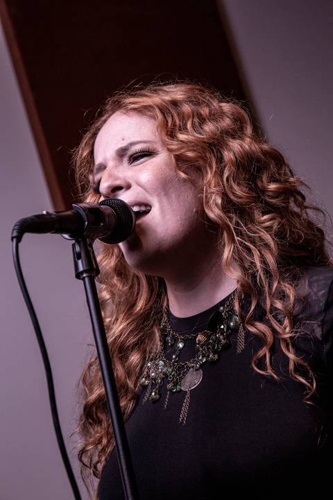 Plano fechado no rosto da vocalista Agnes Gomes, cantando quase perfilada no microfone, com paleta de cor avermelhada e foto levemente inclinada. (Banda Madressilva, Ensaio Musical, Fotos Bruno Arita)'