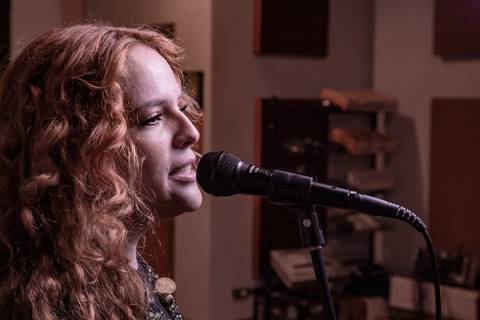 Plano fechado no rosto da vocalista Agnes Gomes, totalmente perfilada e cantando no microfone, com regra dos terços e paleta avermelhada. (Banda Madressilva, Ensaio Musical, Fotos Bruno Arita)'