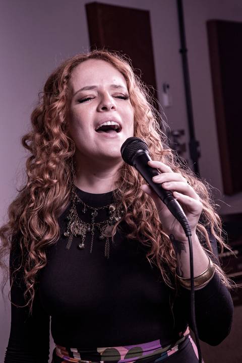 Vocalista Agnes Gomes cantando de frente e segurando o microfone, com a cabeça levemente erguida e plano fechado, com paleta de cor avermelhada. (Banda Madressilva, Ensaio Musical, Fotos Bruno Arita)'