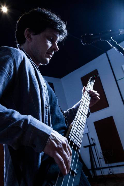 Plano contra-plongée do baixista Marco Augusto tocando, com paleta azulada e alto contraste de luz, gerando impacto. (Banda Madressilva, Ensaio Musical, Fotos Bruno Arita)'
