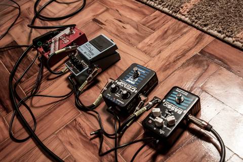 Detalhe dos pedais de efeito do baixista, em sequência, sobre piso de tacos de madeira, com baixa saturação e tom amarelado. (Banda Madressilva, Ensaio Musical, Fotos Bruno Arita)'