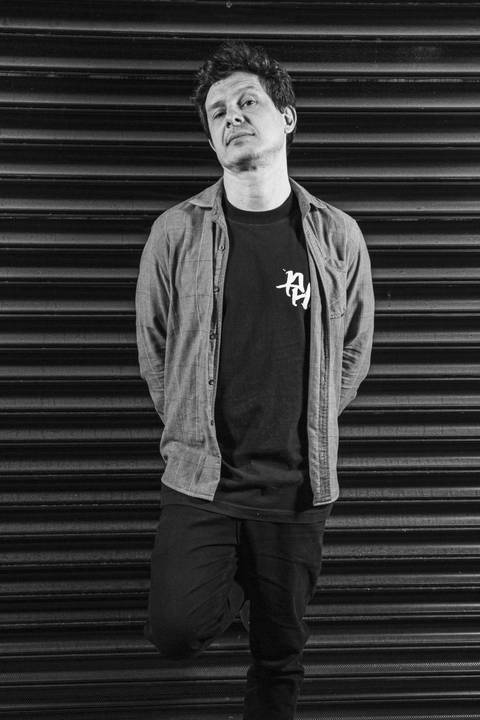 Retrato preto e branco do baixista Marco Augusto encostado no portão preto texturizado, com olhar divertido e curioso para ensaio. (Banda Madressilva, Ensaio Musical, Fotos Bruno Arita)'